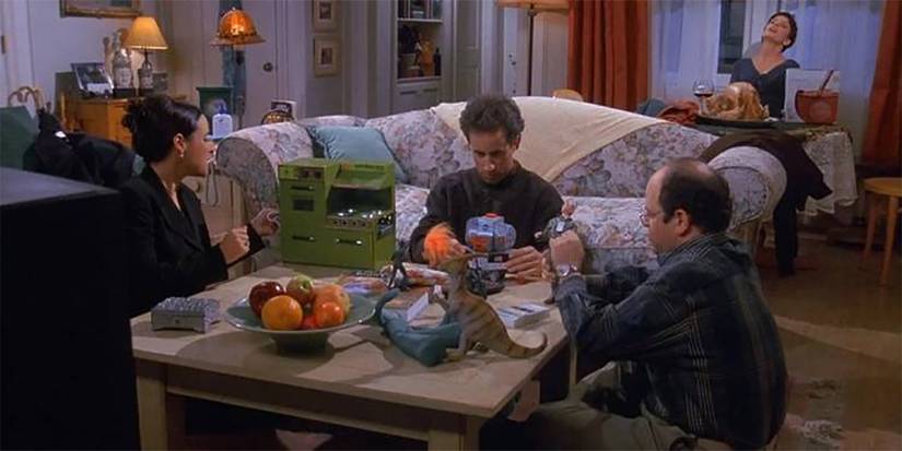 Jerry, Elaine, and George sitting at a table with toys, a woman passed out in the background in Seinfeld.