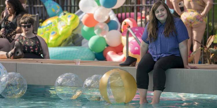 Annie (Aidy Bryant) sitting on the edge of a pool, with her feet in the water, and balloons everywhere in 'Shrill'
