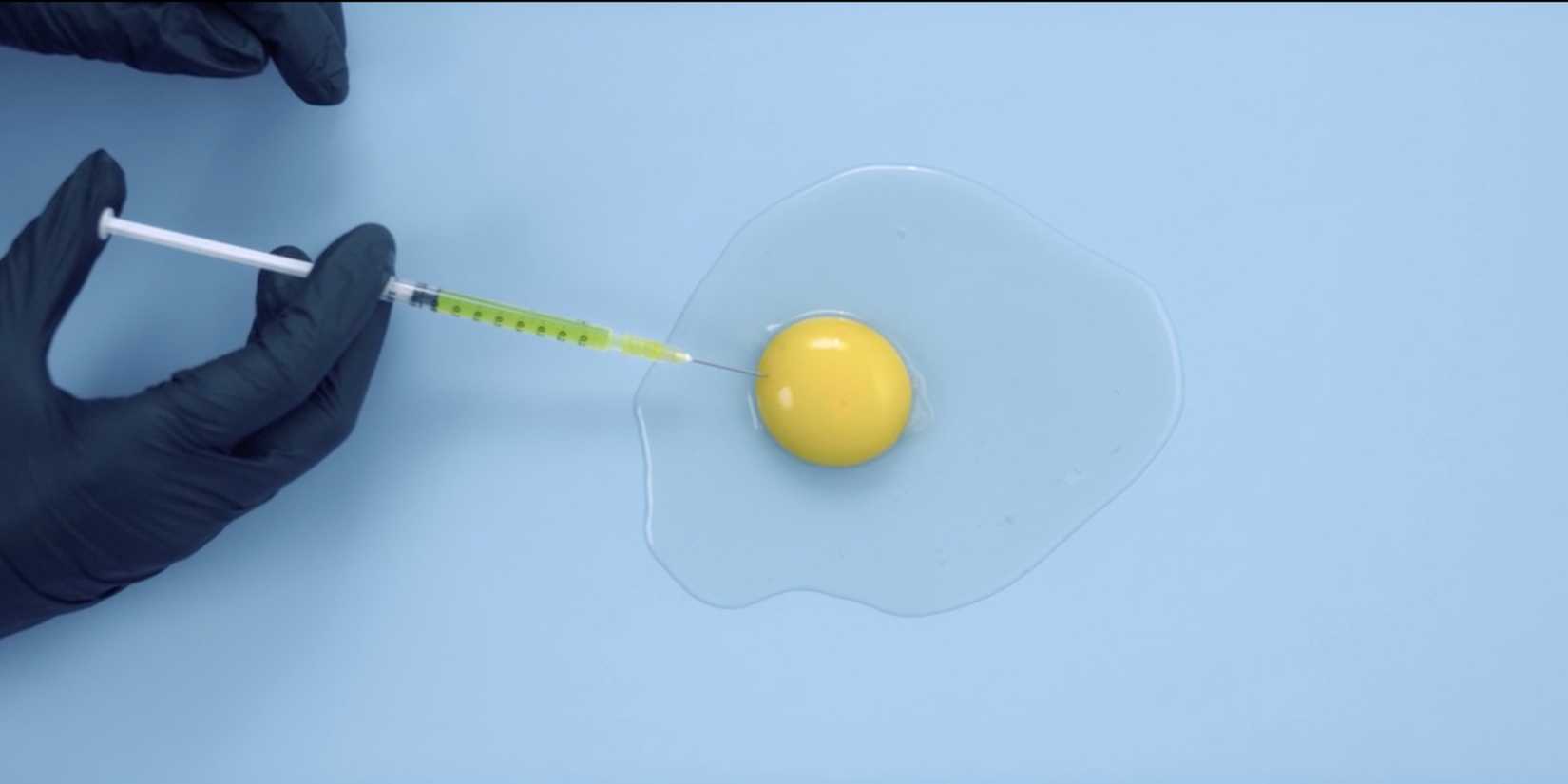 A hand injecting a raw egg yolk with a green liquid in The Substance