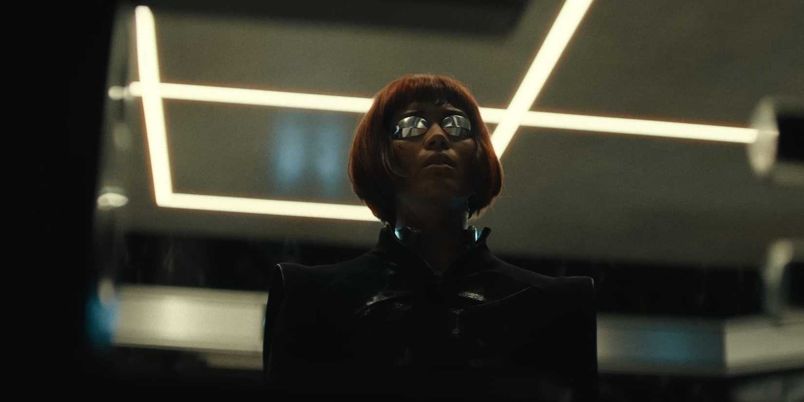 Briana Middleton as Molly Millions in Apple TV's Neuromancer. She is seen at a slight angle from below, partially obscured in shadows with a grid of lights on the ceiling above. She wears a black jacket with shoulder pads and her hair is a short red bob cut.