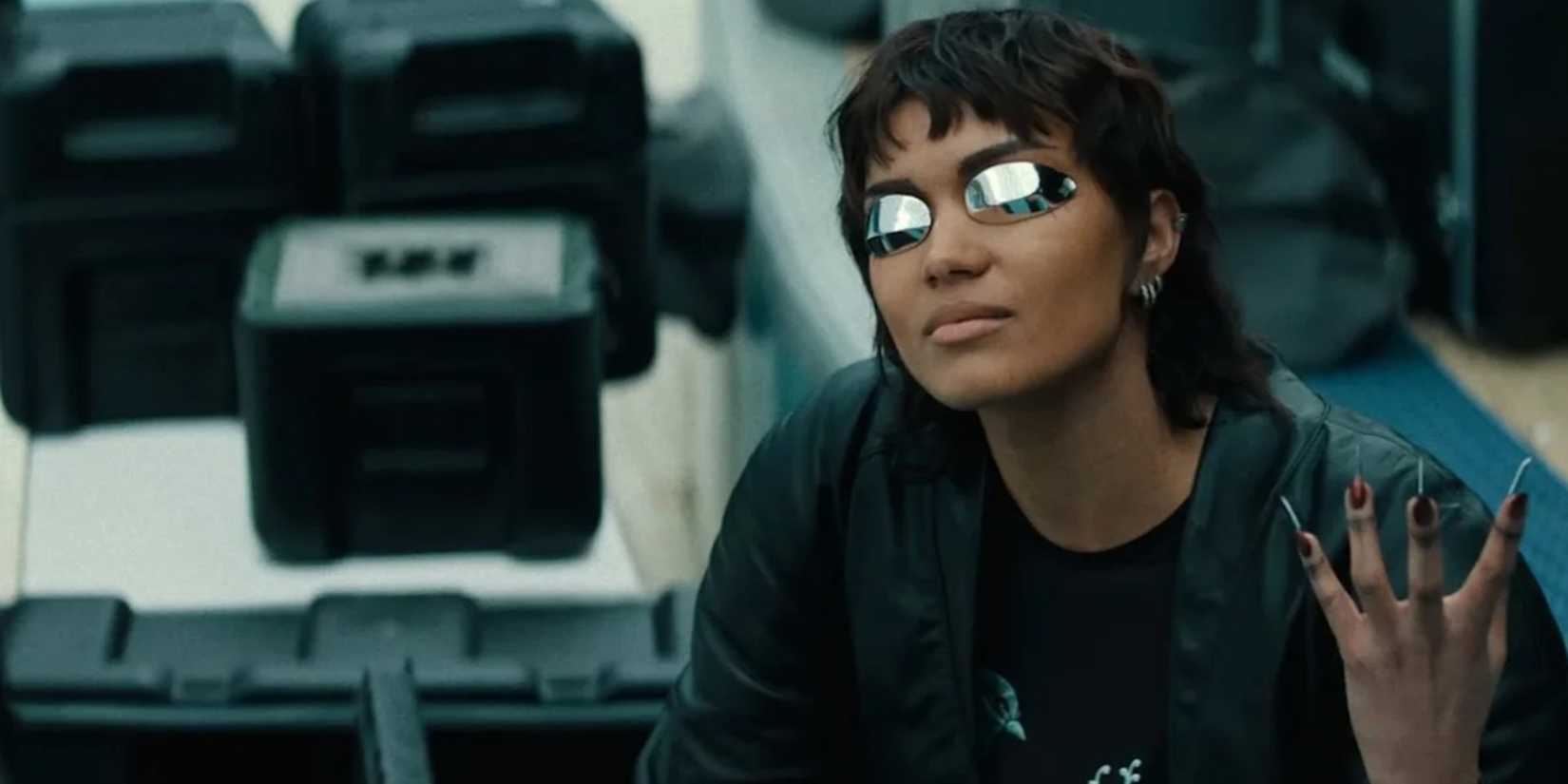 Briana Middleton as Molly Millions in Apple TV's Neuromancer. She sits, surrounded by storage cases, holding up her left hand to display her razor claws. Her expression is neutral.