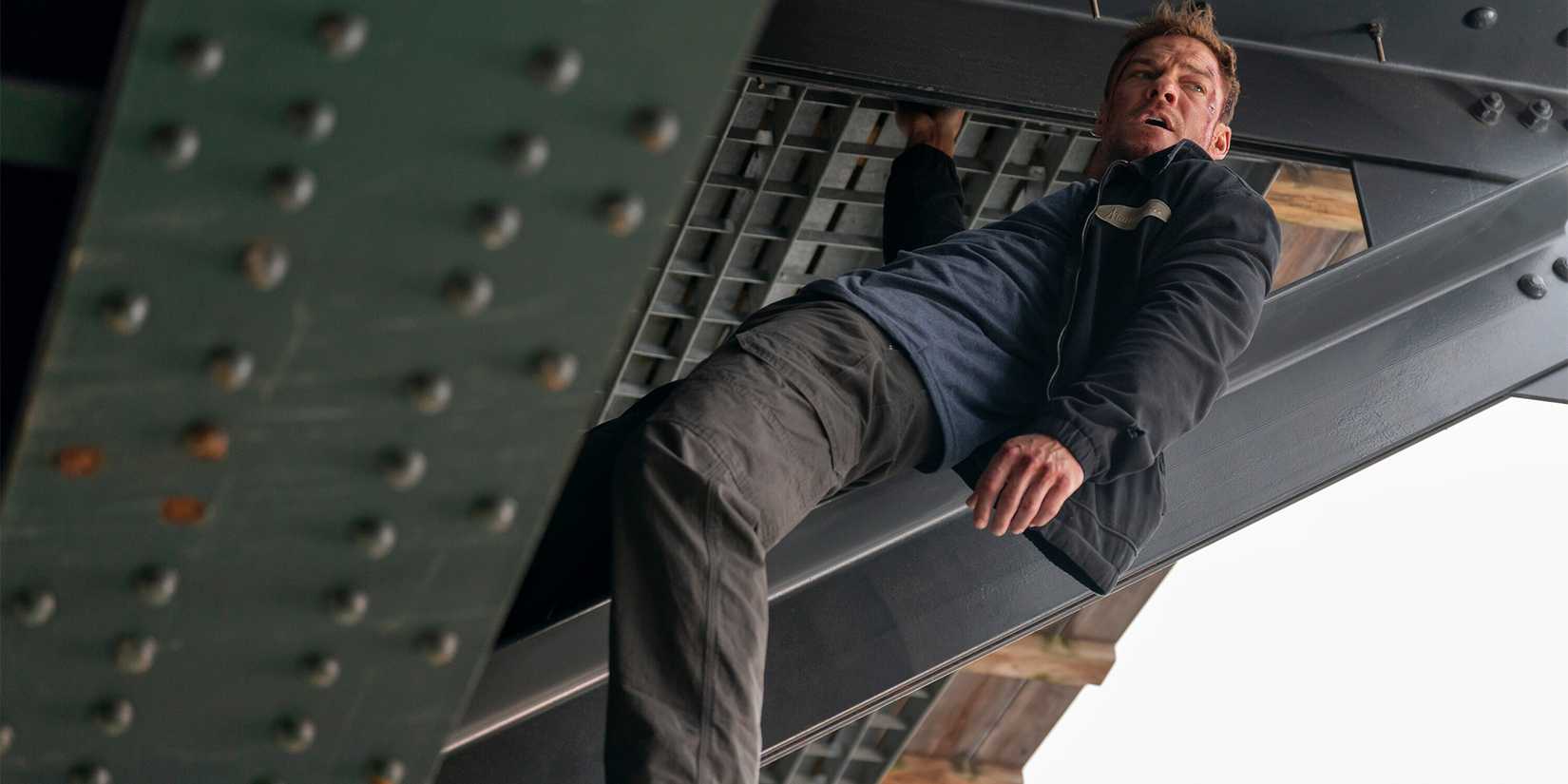 Alan Ritchson as Jack Reacher hanging from the foundation under a bridge