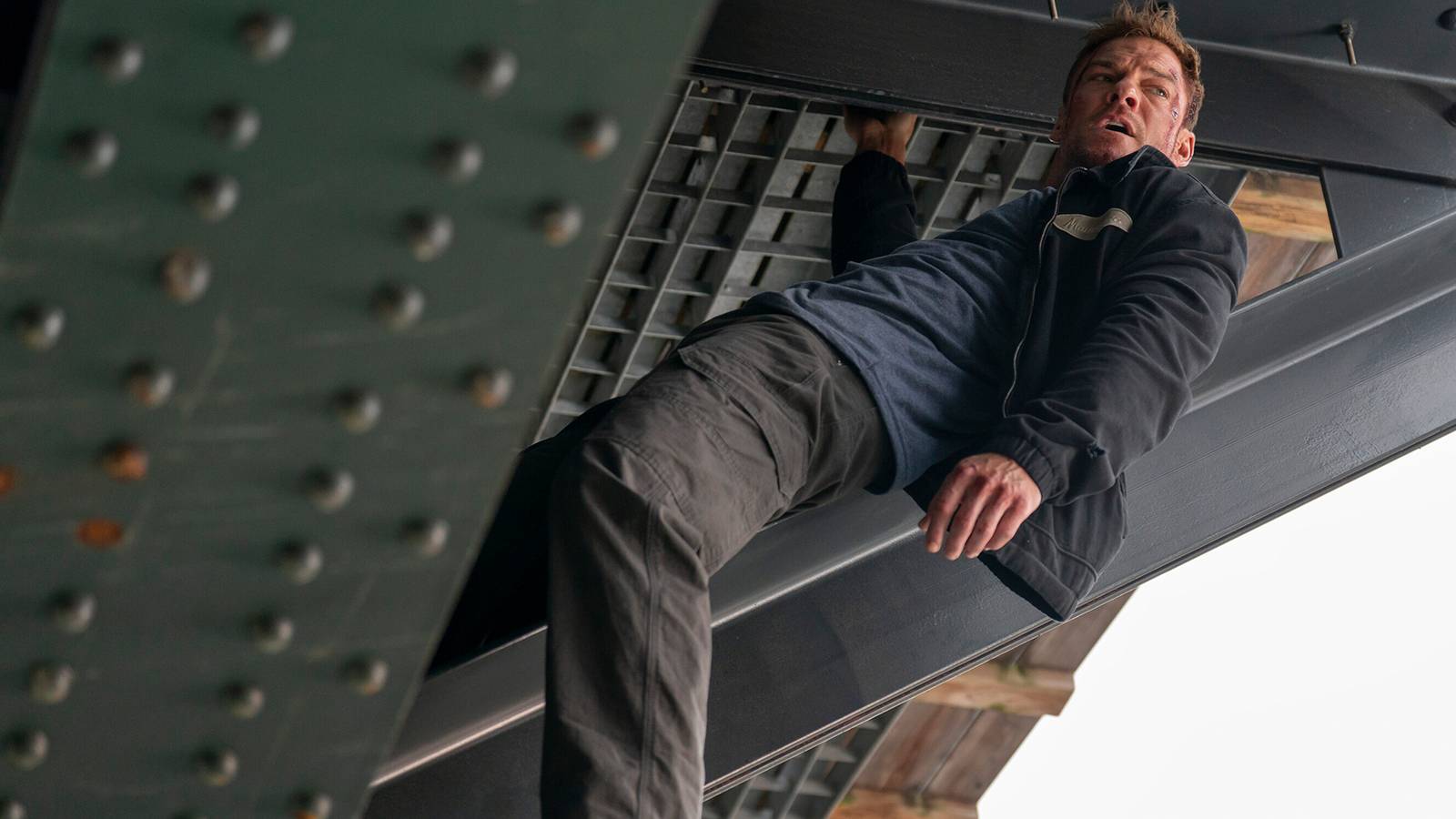 Alan Ritchson as Jack Reacher hanging from the foundation under a bridge