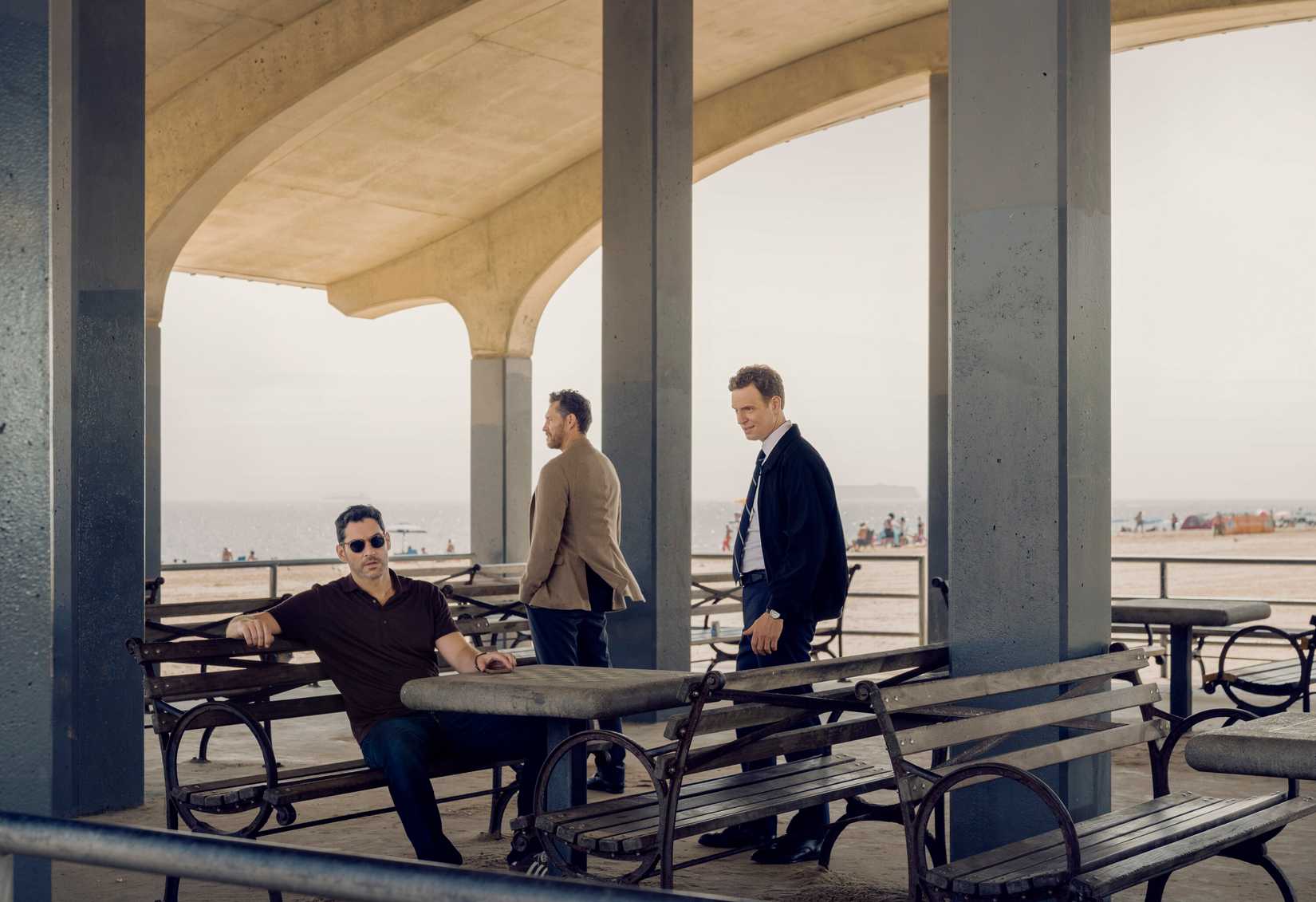 Tom Ellis sitting on a bench by a stone chess board alongside Nick Gehlfuss and Gene Farber in CIA Season 2