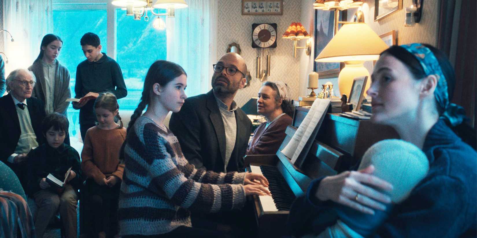 A little girl and her father play piano in a room full of people.