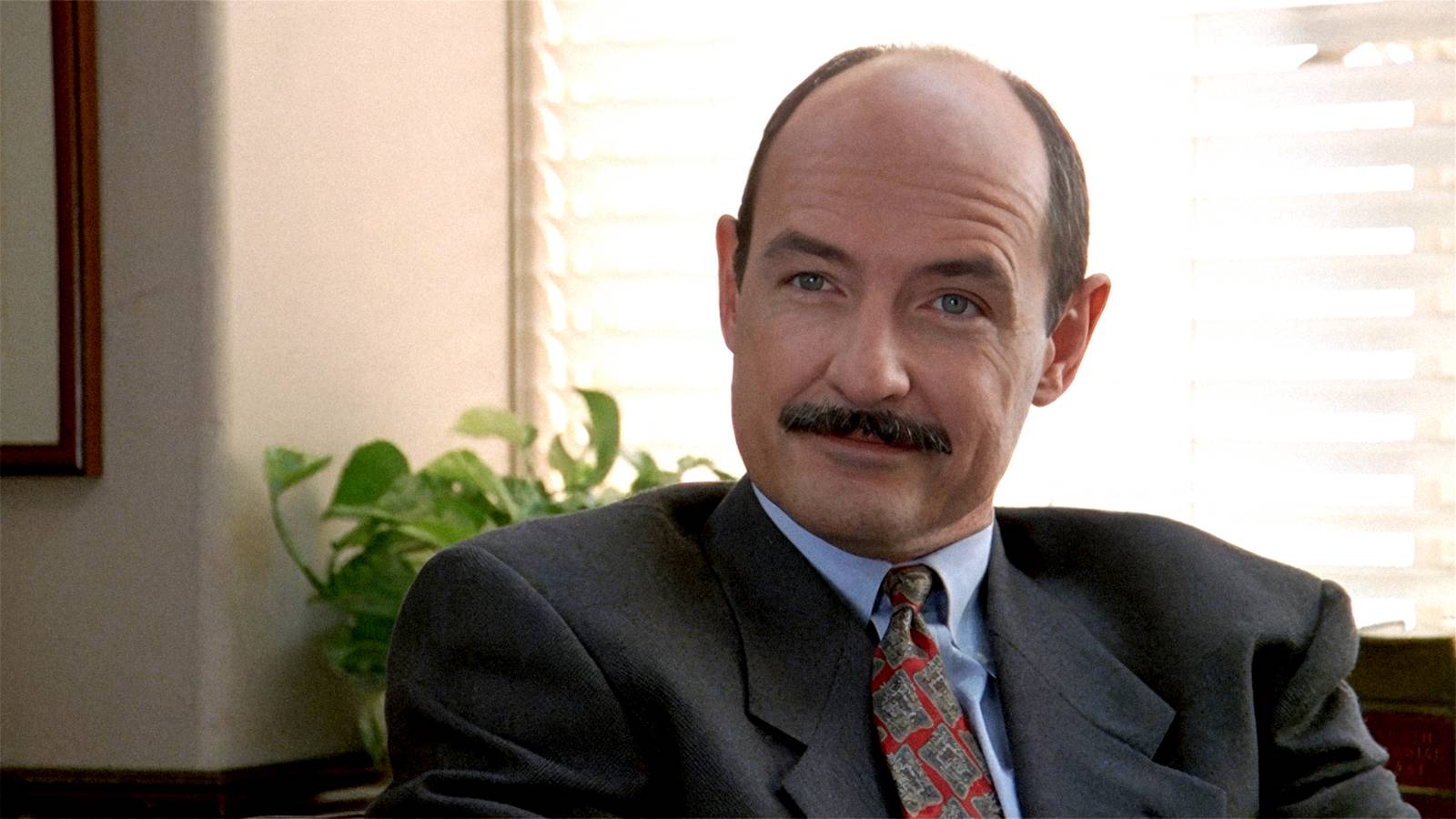 Terry O'Quinn sits at an office desk and smirks in Primal Fear