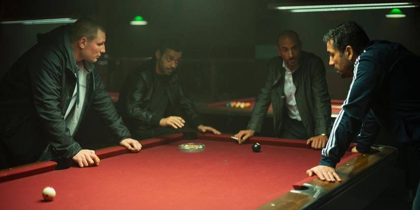 Four men in dark jackets lean over a red pool table talking under dim lighting in a pool hall in The Helicopter Heist